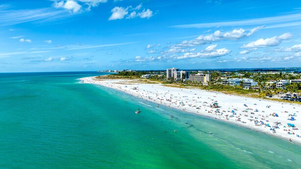 Een strand met helder turquoise water, wit zand en een rij hotels langs de kust.