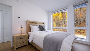 Une chambre avec une tête de lit en bois, une table de chevet avec une lampe, une grande fenêtre donnant sur le feuillage automnal et une porte.