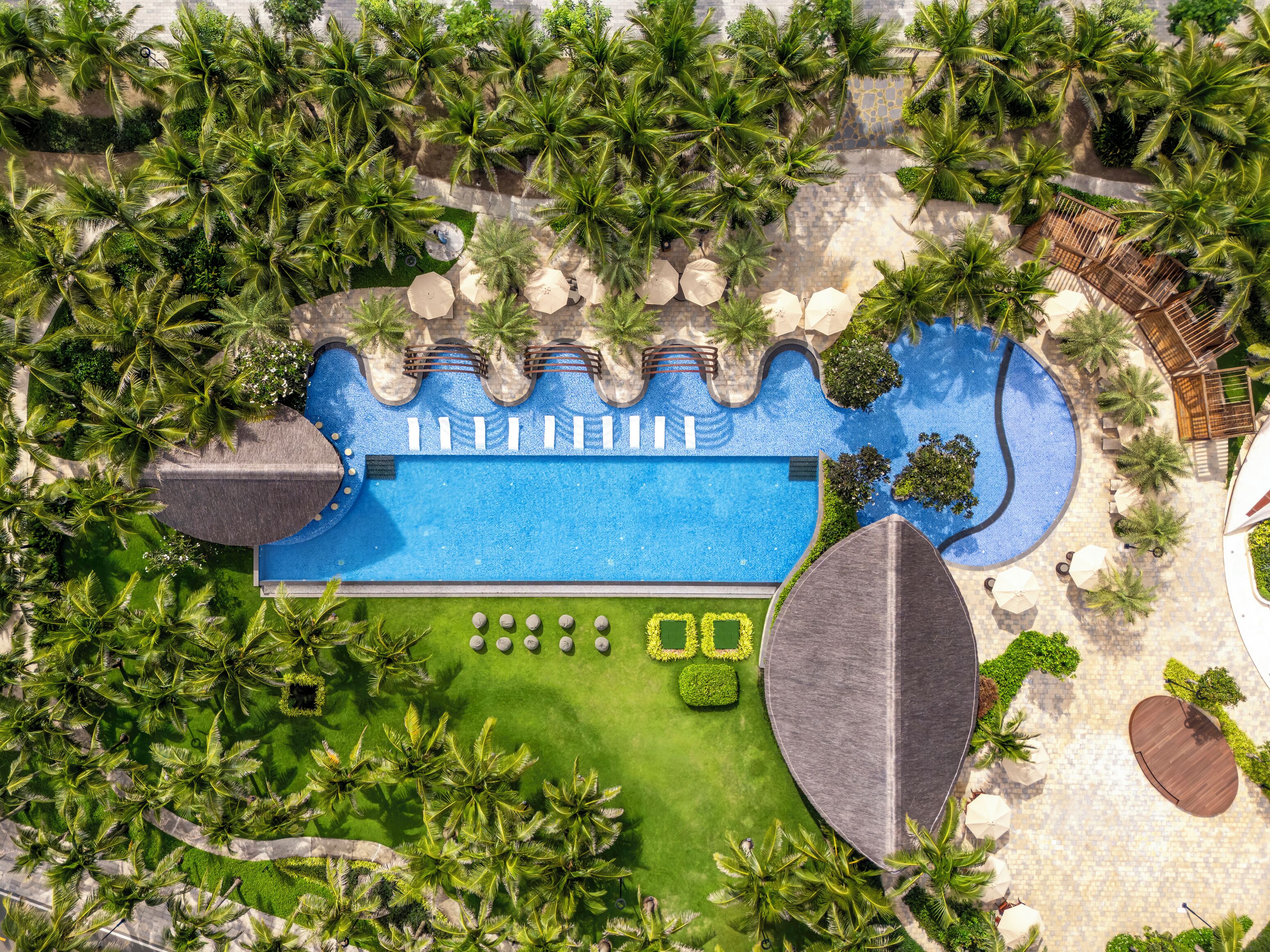 An aerial view of a resort with a central swimming pool, surrounded by palm trees and sun loungers.
