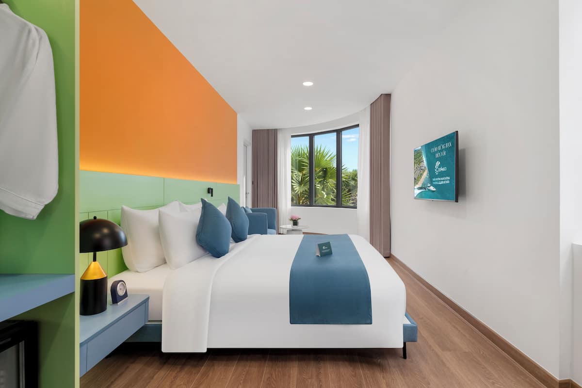 Une chambre d’hôtel moderne avec un grand lit, une table de chevet, une lampe, une horloge, une fenêtre donnant sur des palmiers et un téléviseur fixé au mur.