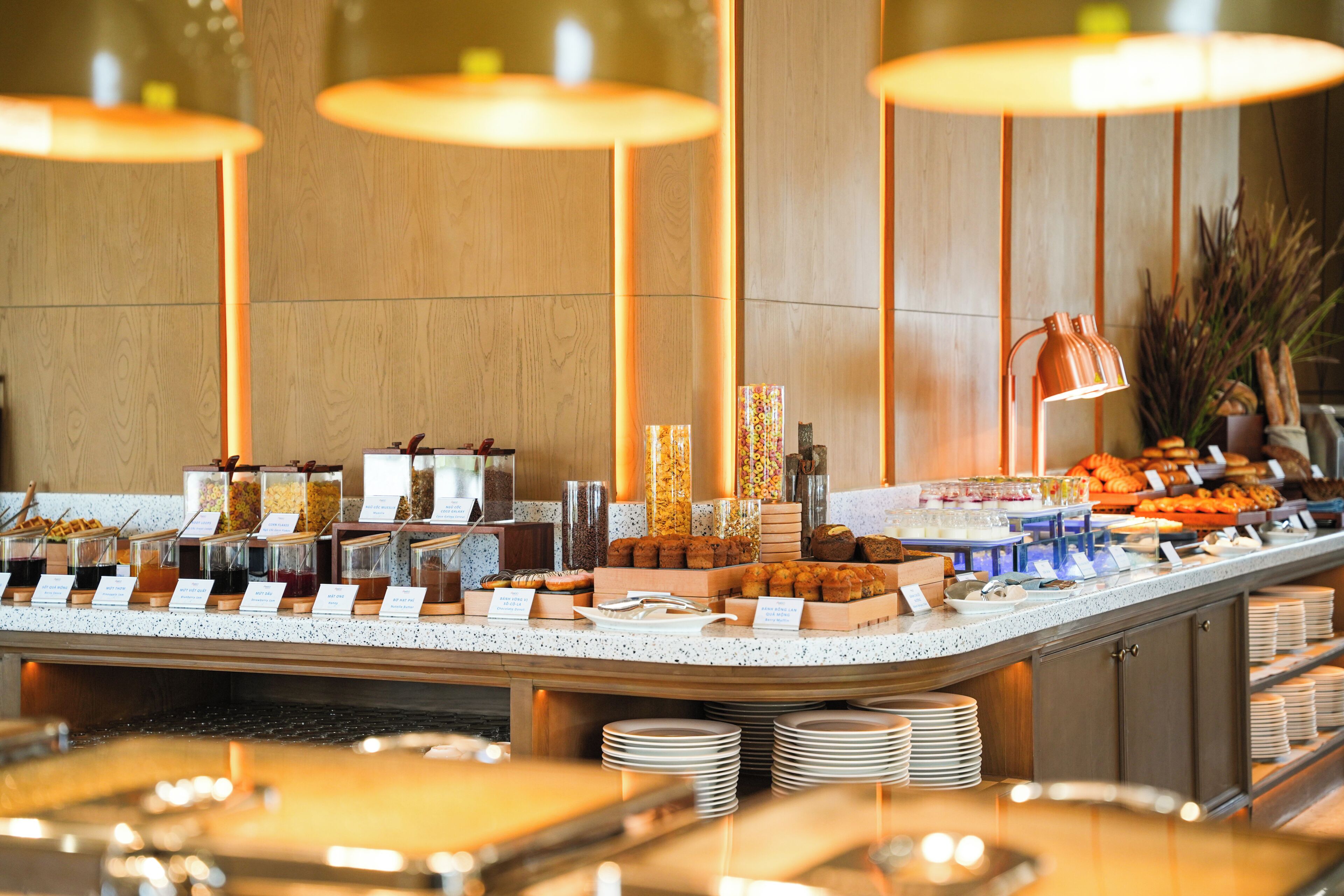 A buffet with various food items and containers, a stack of plates, and a lamp.