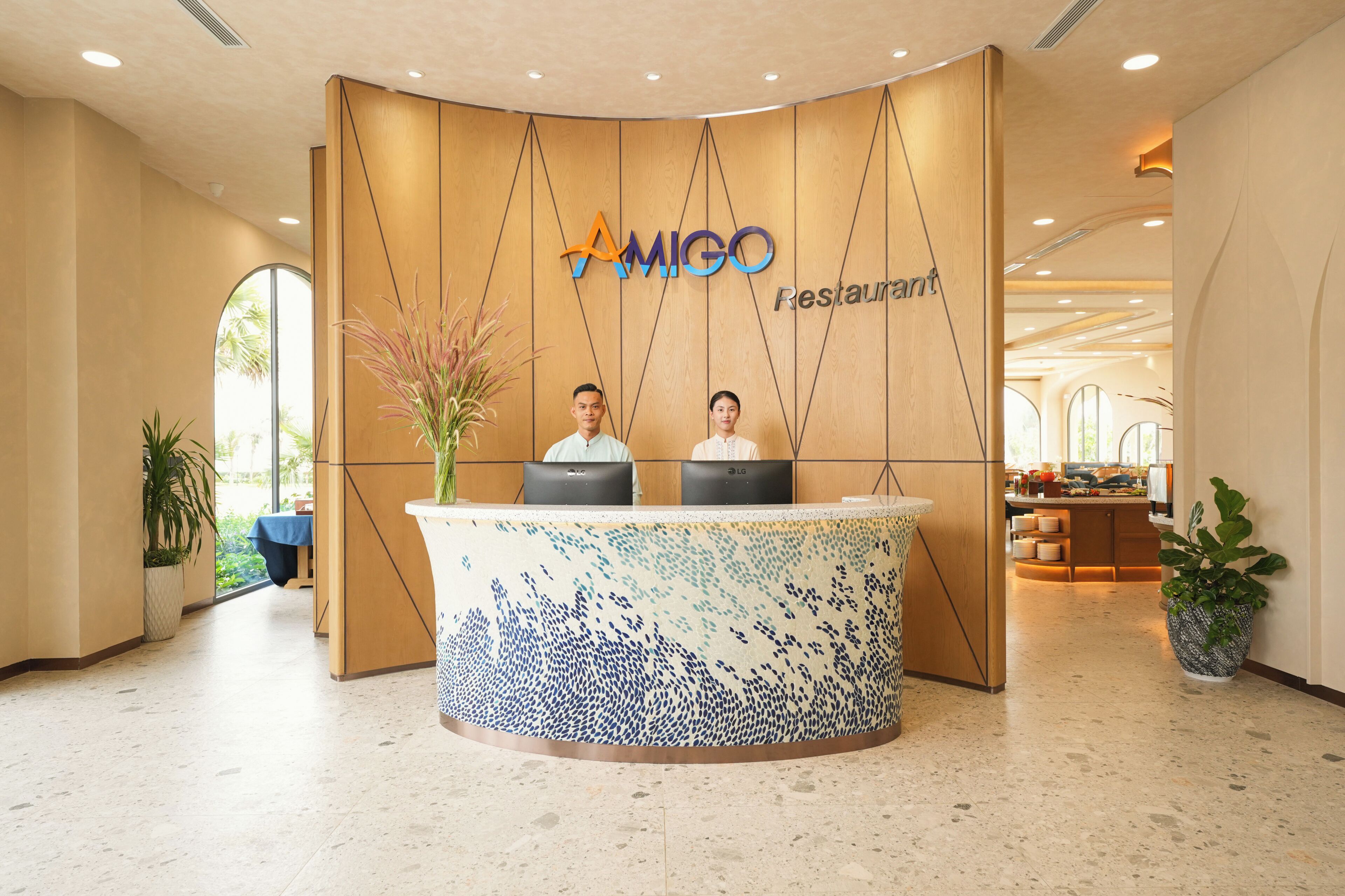 Un mostrador de recepción con un logotipo, dos personas sentadas y una planta en maceta.