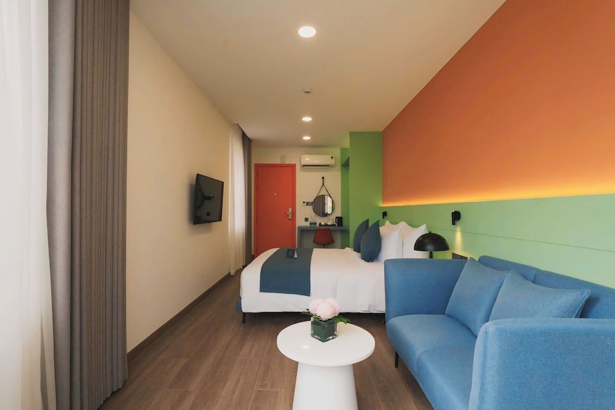 Une chambre d’hôtel moderne avec un lit, un canapé bleu, une petite table blanche ornée d’une fleur, et une porte rouge en arrière-plan.