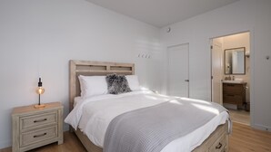 A bedroom with a bed, bedside table, and a nightstand. There is a door leading to a bathroom. - Panorama 1249: Majestic River View, Spacious Patio (Petite-Rivière-Saint-François)