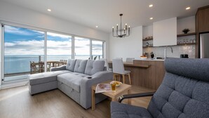 A modern living room with a large gray sectional sofa, a dining area with wooden chairs, a kitchen with white cabinets, and a view of the sea through large windows. - Panorama 1251: Private & Luxury, River Sunsets (Petite-Rivière-Saint-François)