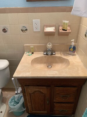 A bathroom with a beige sink, a wooden vanity, a toilet, and a trash can. There are two soap dispensers on the wall and a hand soap bottle on the sink. - Pink Flamingo - Pet Friendly, Beach Side, Wi-Fi (Norfolk)