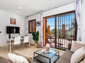 Ein Wohnzimmer mit Sofa, Couchtisch, Fernseher, Essbereich und einer Glastür zum Außenbereich.