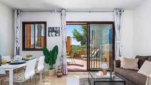Ein modernes Wohnzimmer mit einem Glastisch, einer Couch, einem Essbereich mit Stühlen und einer Glasschiebetür, die auf eine Terrasse im Freien führt.