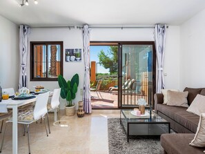 Ein modernes Wohnzimmer mit einem Glastisch, einer Couch, einem Essbereich mit Stühlen und einer Glasschiebetür, die auf eine Terrasse im Freien führt.