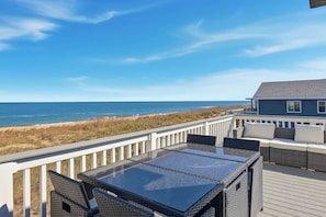 A deck with a glass-top table and wicker chairs, overlooking a beach and ocean.