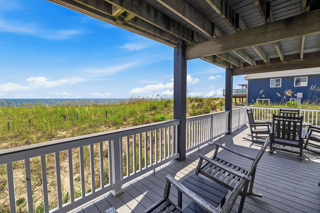 A wooden deck with outdoor seating, overlooking a beach and a building with a blue facade.