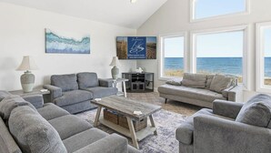 A spacious living room with grey sofas, a wooden coffee table, and a large window overlooking the sea. - Anthem of the Sun | Oceanfront | Private Pool and Hot Tub (Virginia Beach)