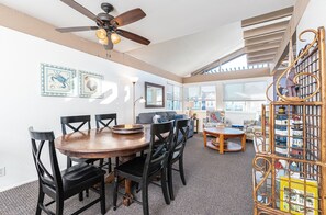 A spacious living room with a round wooden dining table, black chairs, a ceiling fan, and a decorative bookshelf. - Charming 6BR Coastal Escape with Oceanview Patio, Steps from the Sand! (Newport Beach)