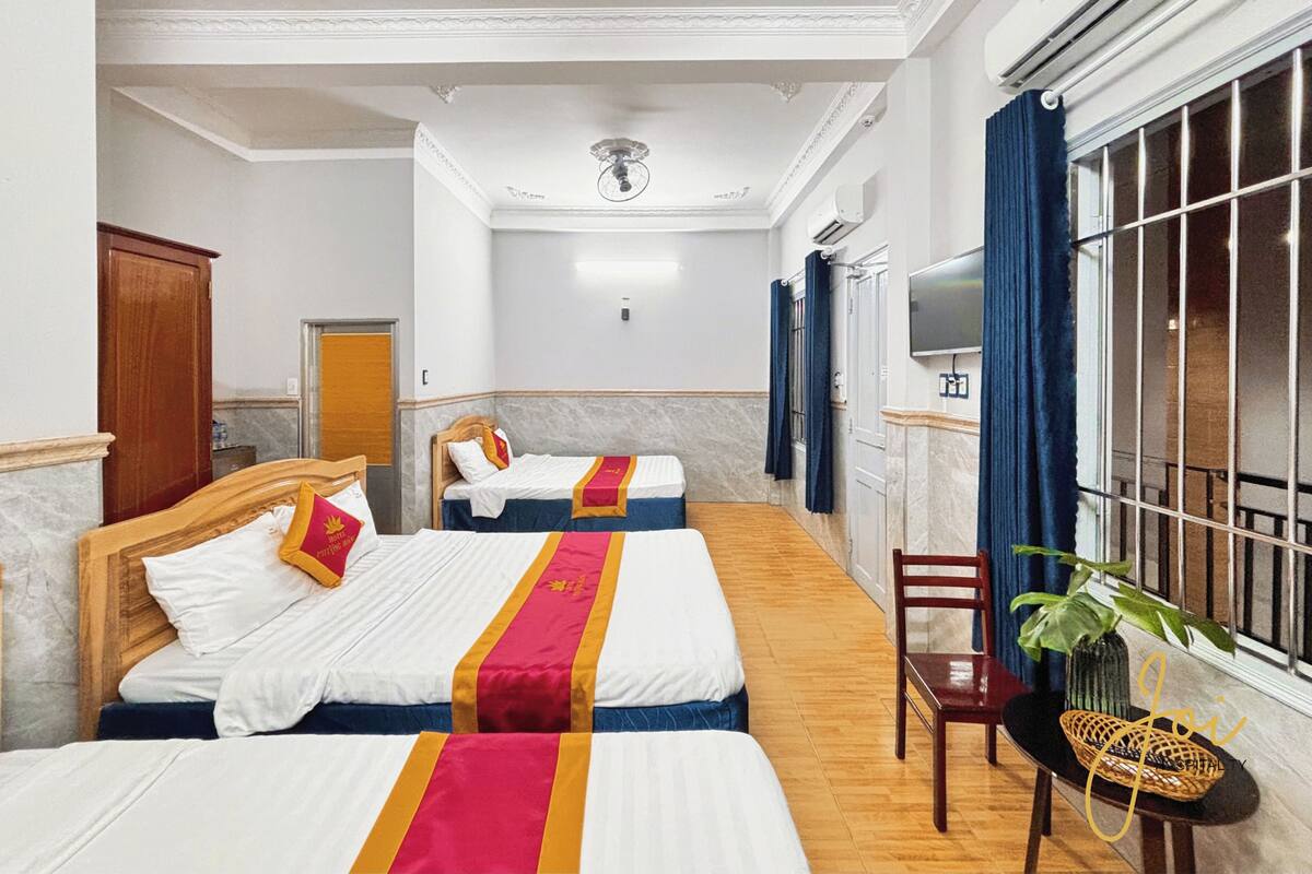 Ein Hotelzimmer mit zwei Betten, Holzboden, Fernseher, einem Fenster mit Vorhängen und einem kleinen Tisch mit Pflanze.