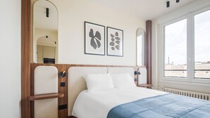 Een slaapkamer met een bed, een houten hoofdeinde, een groot raam en twee ingelijste foto’s aan de muur.