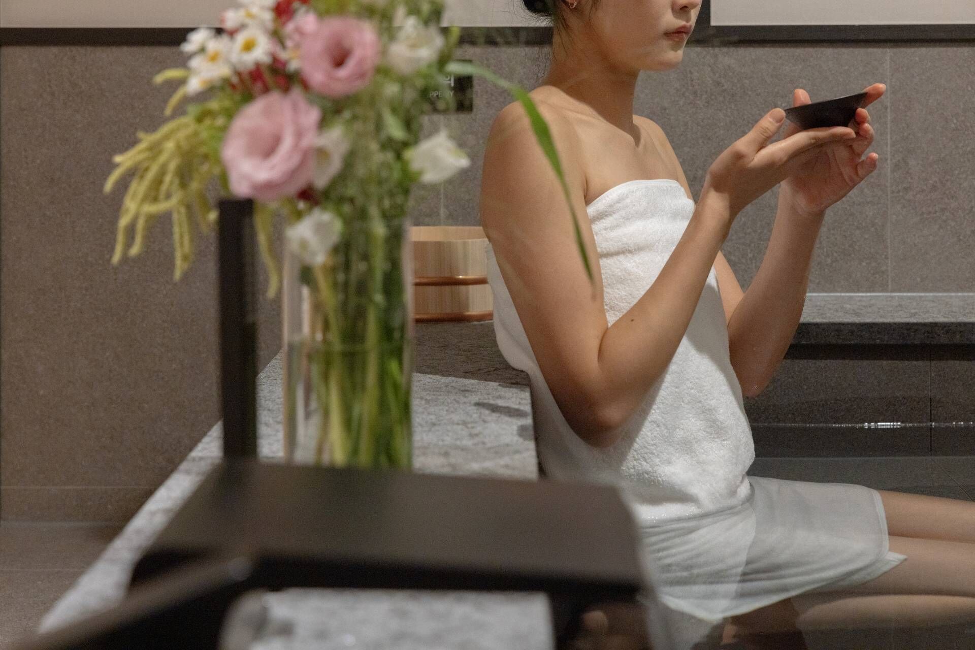 A person in a white towel sitting on a bench in a room with a marble floor and a wall-mounted mirror.