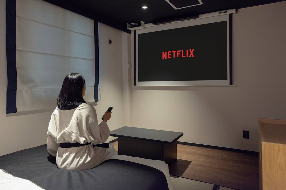 Eine Person sitzt auf einem Bett in einem Raum, in dem ein großer Bildschirm das Wort „NETFLIX“ in roten Buchstaben zeigt.