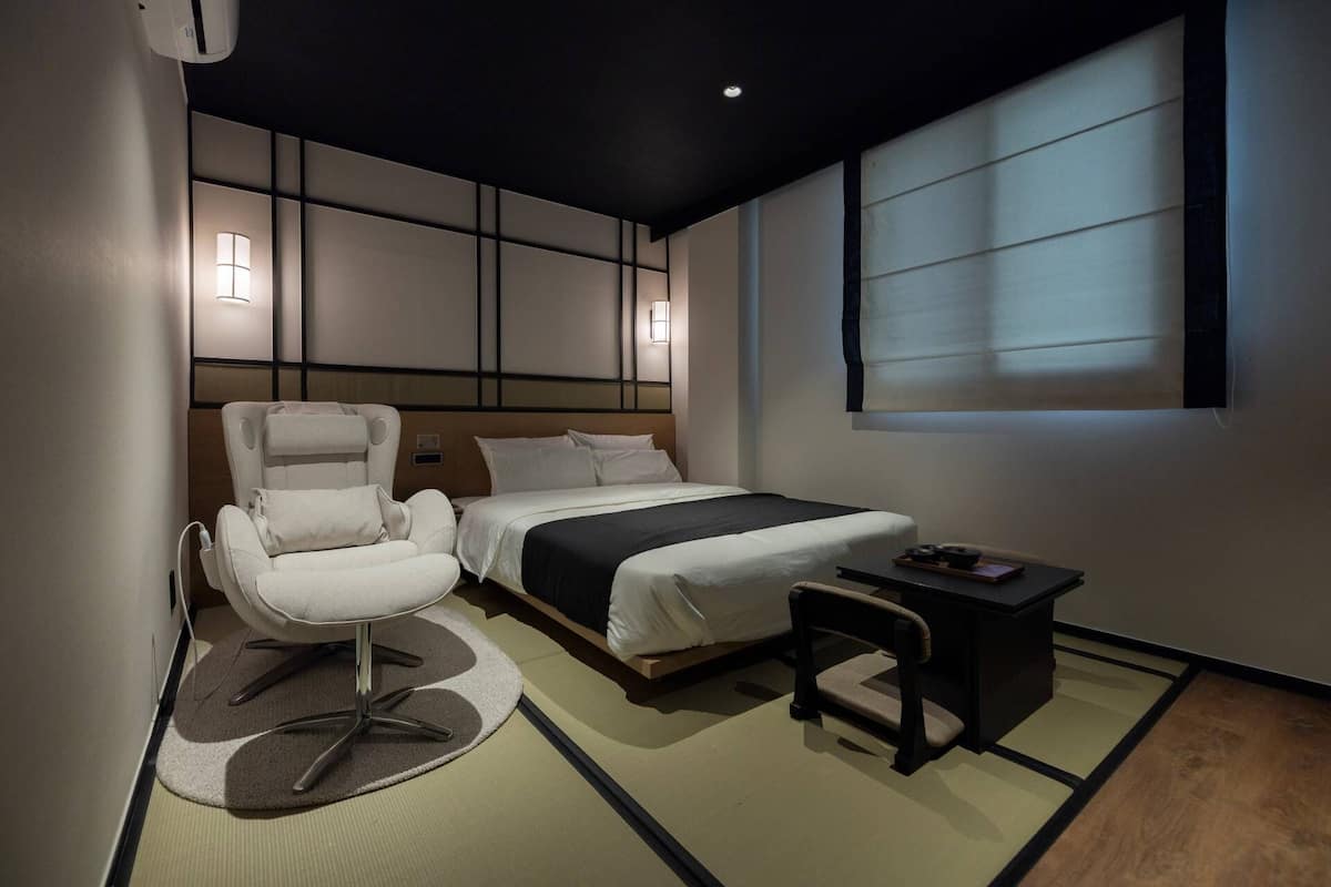Ein modernes Hotelzimmer mit einem Bett, einem Massagesessel, einem kleinen Tisch und einem Fenster mit Jalousien.