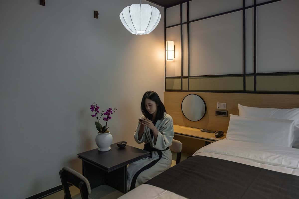 Ein Hotelzimmer mit einem Bett, einem Nachttisch mit Blumenvase, einem Schreibtisch und einem Stuhl.