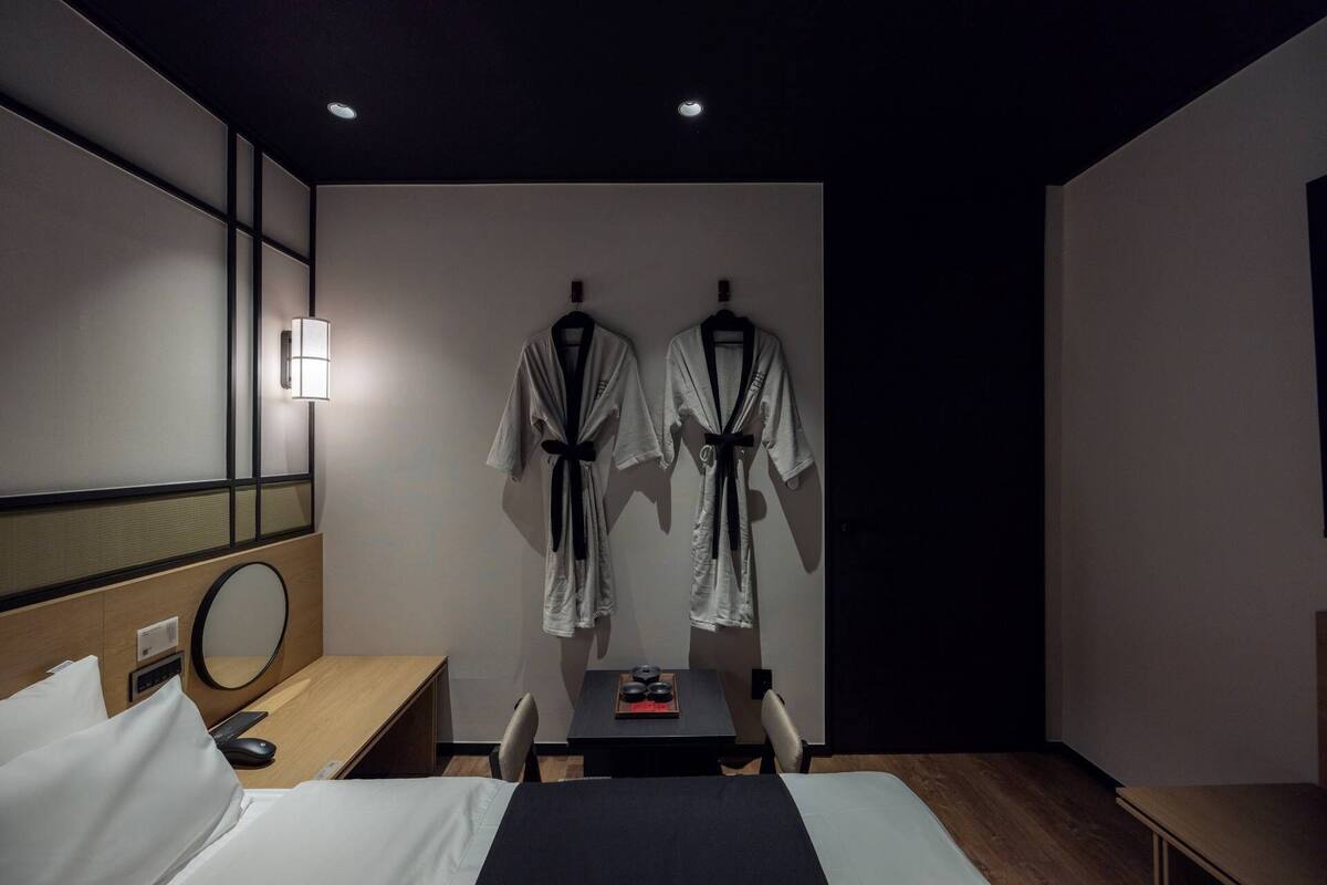 Ein Hotelzimmer mit einem Bett, einem Schreibtisch, einem Stuhl, einem Spiegel und zwei Bademänteln, die an der Wand hängen.