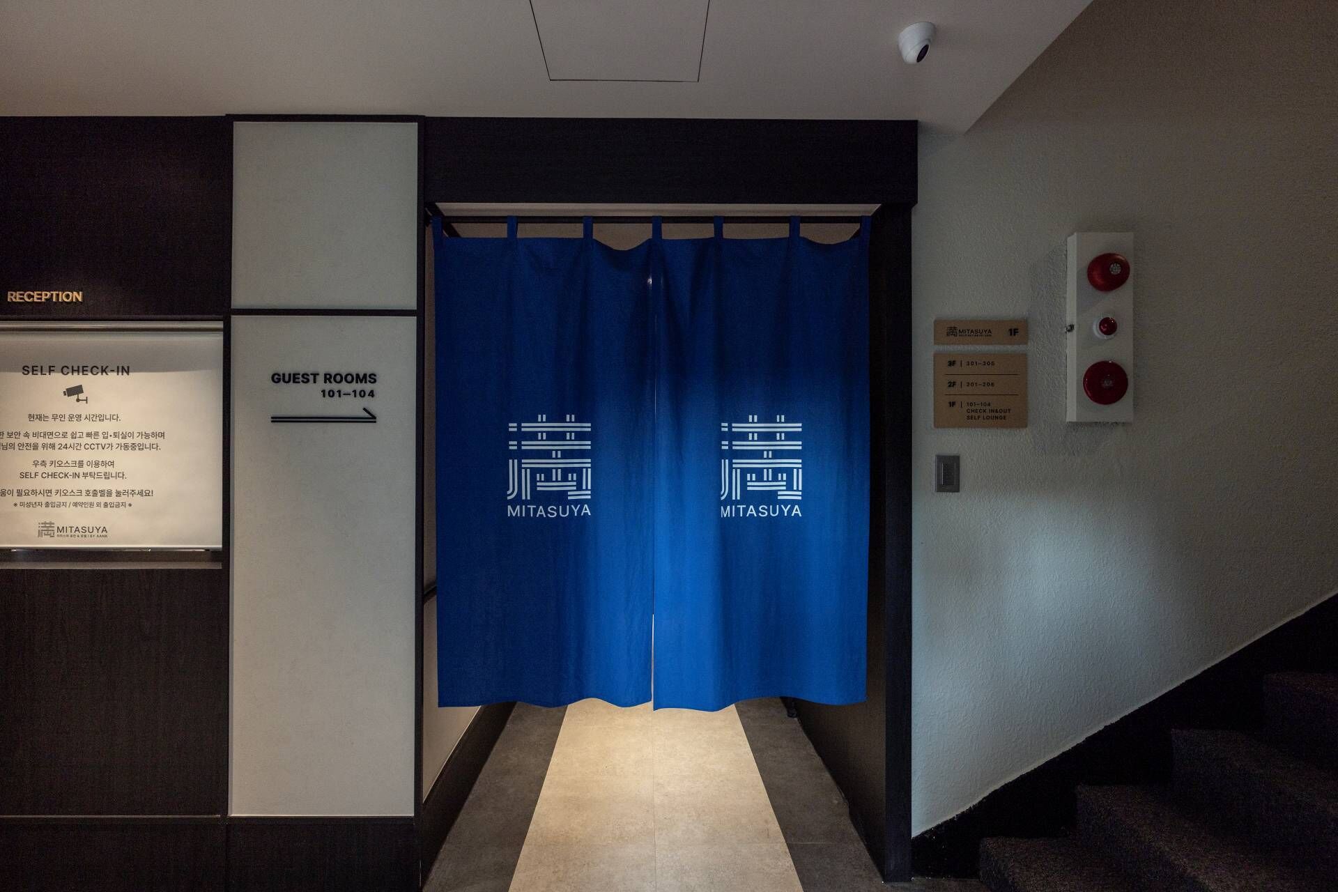 Zona de recepción de un hotel con un mostrador de autopregistro, habitaciones de huéspedes señalizadas con los números 101 a 104, y una cortina azul con el nombre del hotel.