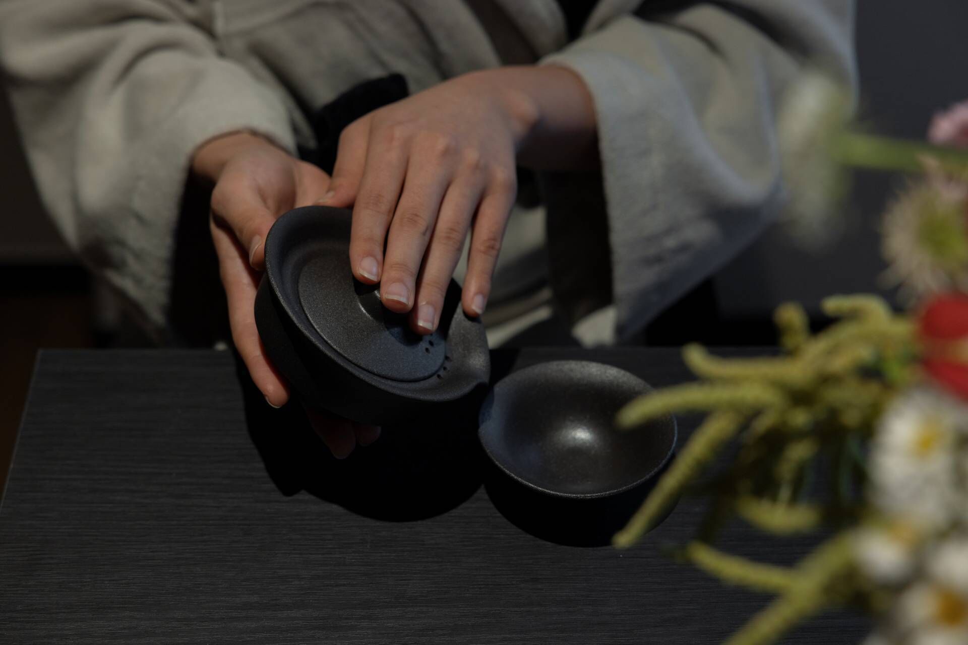 A person holding a black object, possibly a container, with a small bowl beside it on a dark surface.