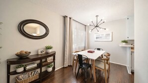 Un coin repas avec une table blanche, des chaises en bois, une console sombre agrémentée d’objets décoratifs, un miroir et un luminaire moderne.