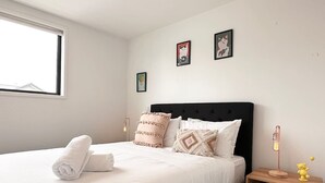 Een net opgemaakt bed met een opgerolde witte handdoek, een zwart hoofdbord en twee decoratieve kussens. Drie ingelijste foto’s hangen aan de muur, een bedlamp en een klein houten nachtkastje met een teddybeer.