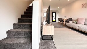 Een moderne woonkamer met een trap, een flatscreen-tv op een houten voetstuk, een bank en een zithoek met stoelen en een tafel.