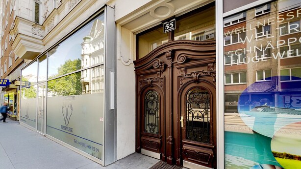 A building facade with a large glass window advertising a wellness center, a wooden ornate door with a brass handle, and a street sign above the door. - Blueground | Josefstadt, washer/dryer, nr Votivkirche (Vienna)