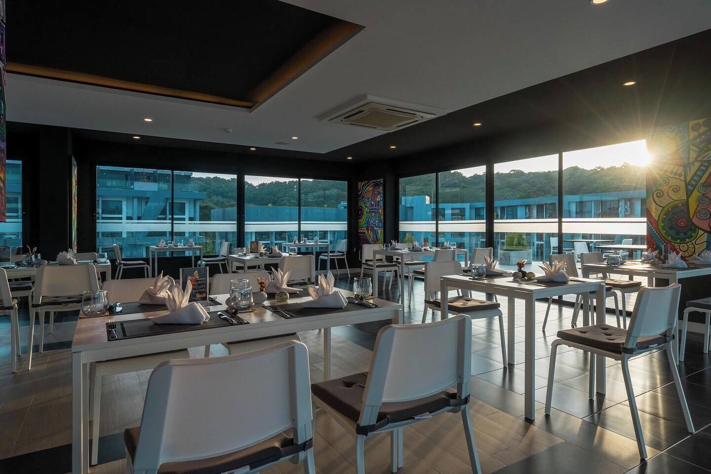 Un'elegante sala da pranzo con tavoli apparecchiati, ampie finestre che offrono vista su edifici e aree verdi, e un soffitto con illuminazione a incasso.
