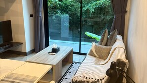 A modern living room with a sofa, coffee table, and TV. There is a large window with curtains, offering a view of greenery outside. - Utopia Loft Aparthotel by Sabai Sands (Rawai)