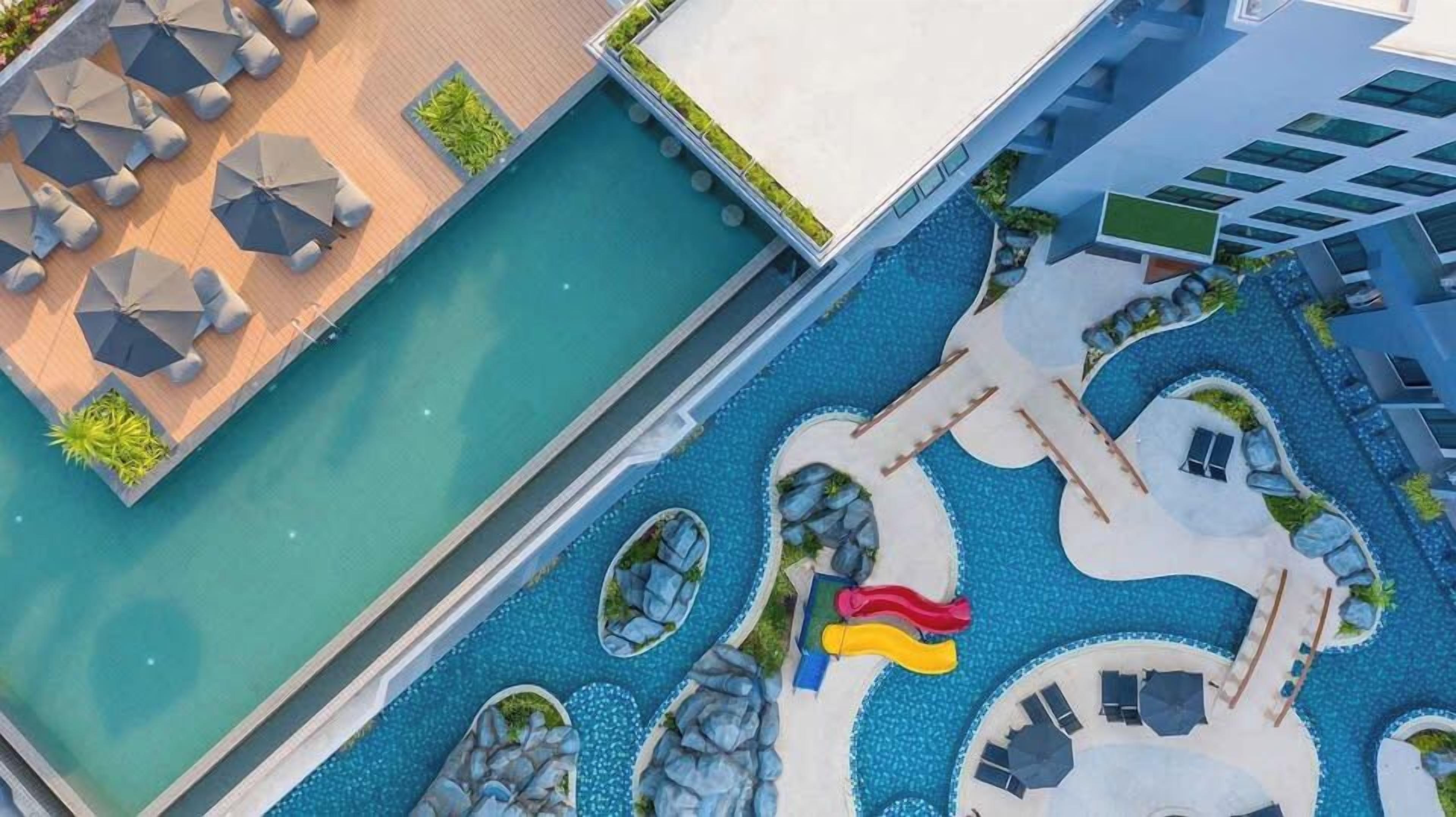 Un área de piscina en la azotea con varias zonas de estar, un tobogán y un camino que lleva a la entrada del edificio.