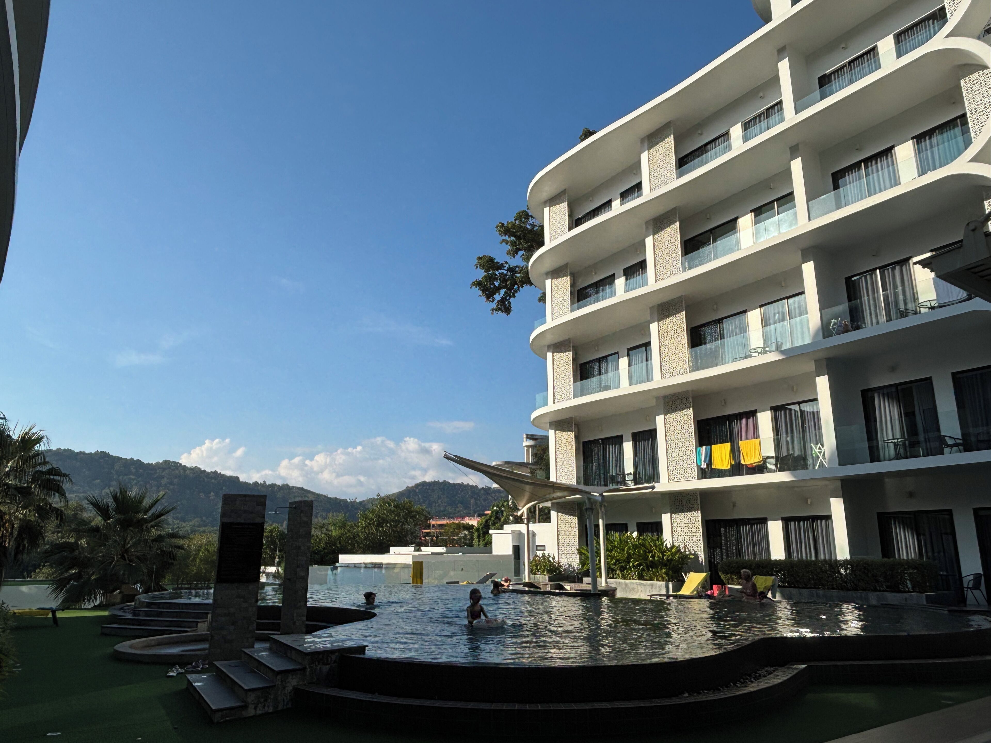 A modern hotel with a pool area, featuring a multi-level structure with balconies and a view of the mountains.