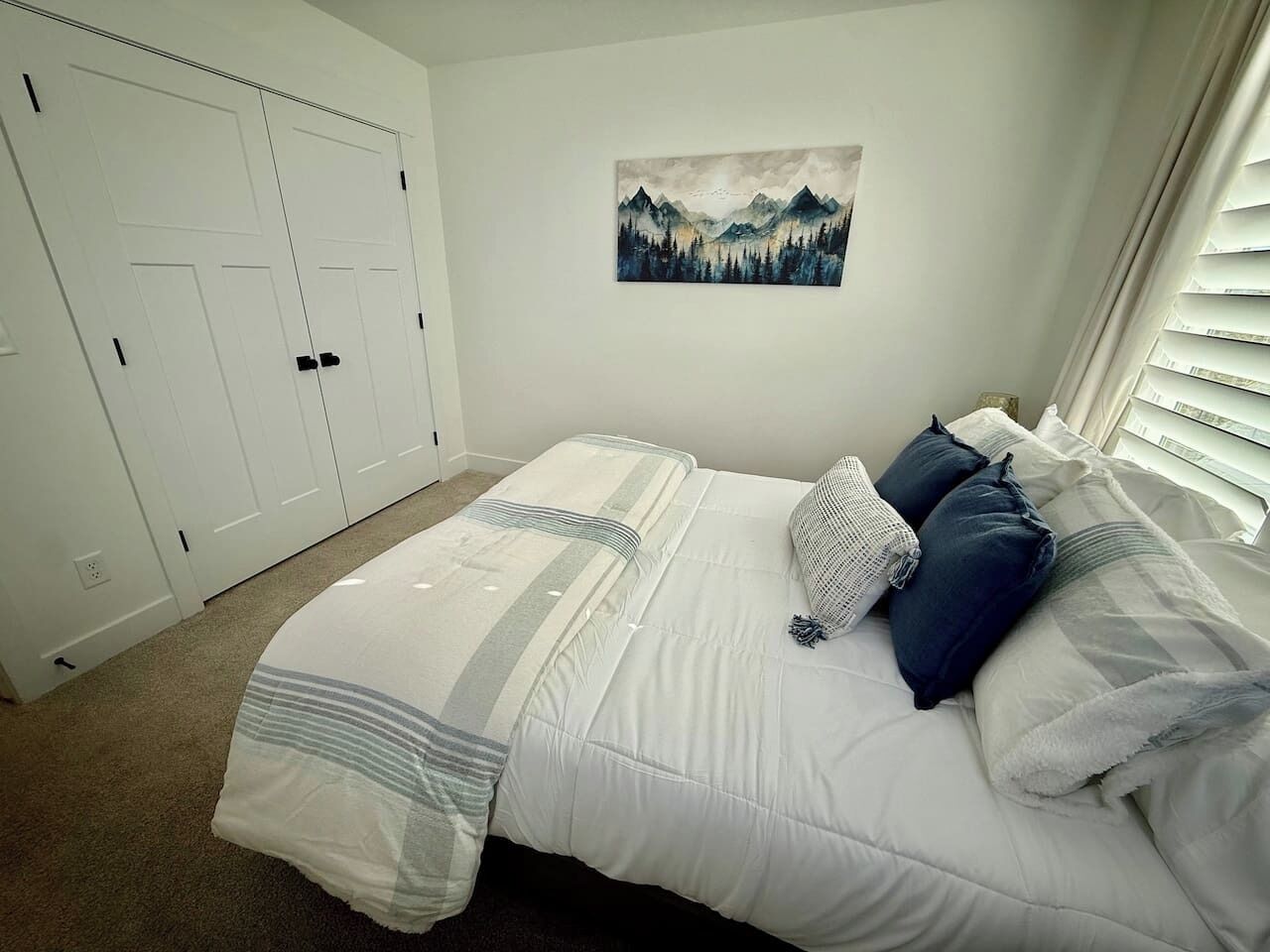 A bedroom with a bed, white walls, a mountain painting, and a closet.