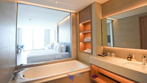 A modern bathroom with a large bathtub, a vanity with a mirror, and a view of a bedroom with a bed and a desk. - OceanView Hạ Long • Large Balcony • TwinBed (Thành phố Hạ Long)