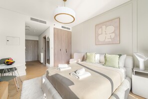 A modern bedroom with a large bed, a bedside table, a chair, and a desk with a bowl of fruit. - Peninsula One - 1312 Studio (Dubai)