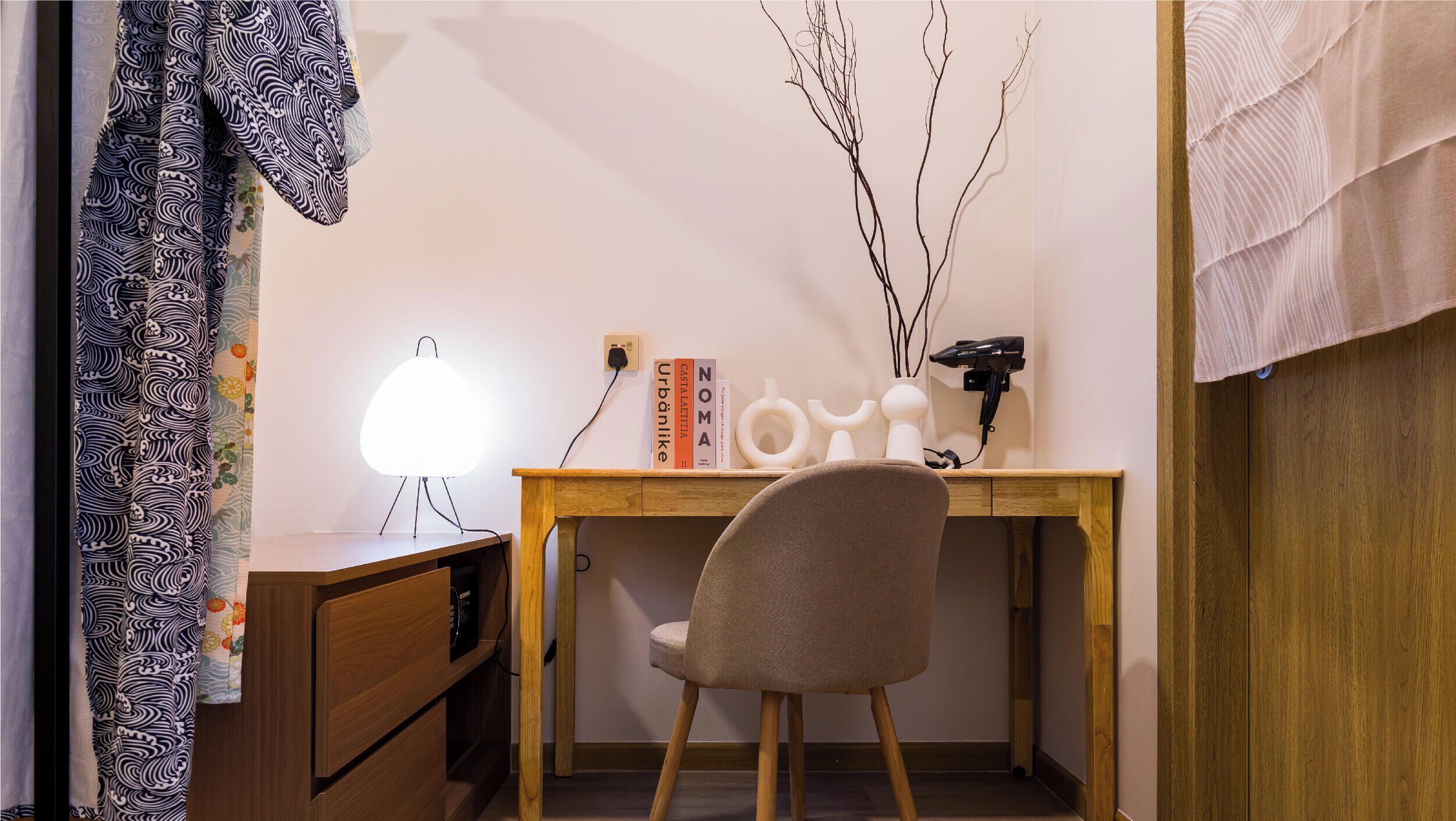 Una habitación con escritorio de madera, una silla, lámpara, libros y secador de pelo.