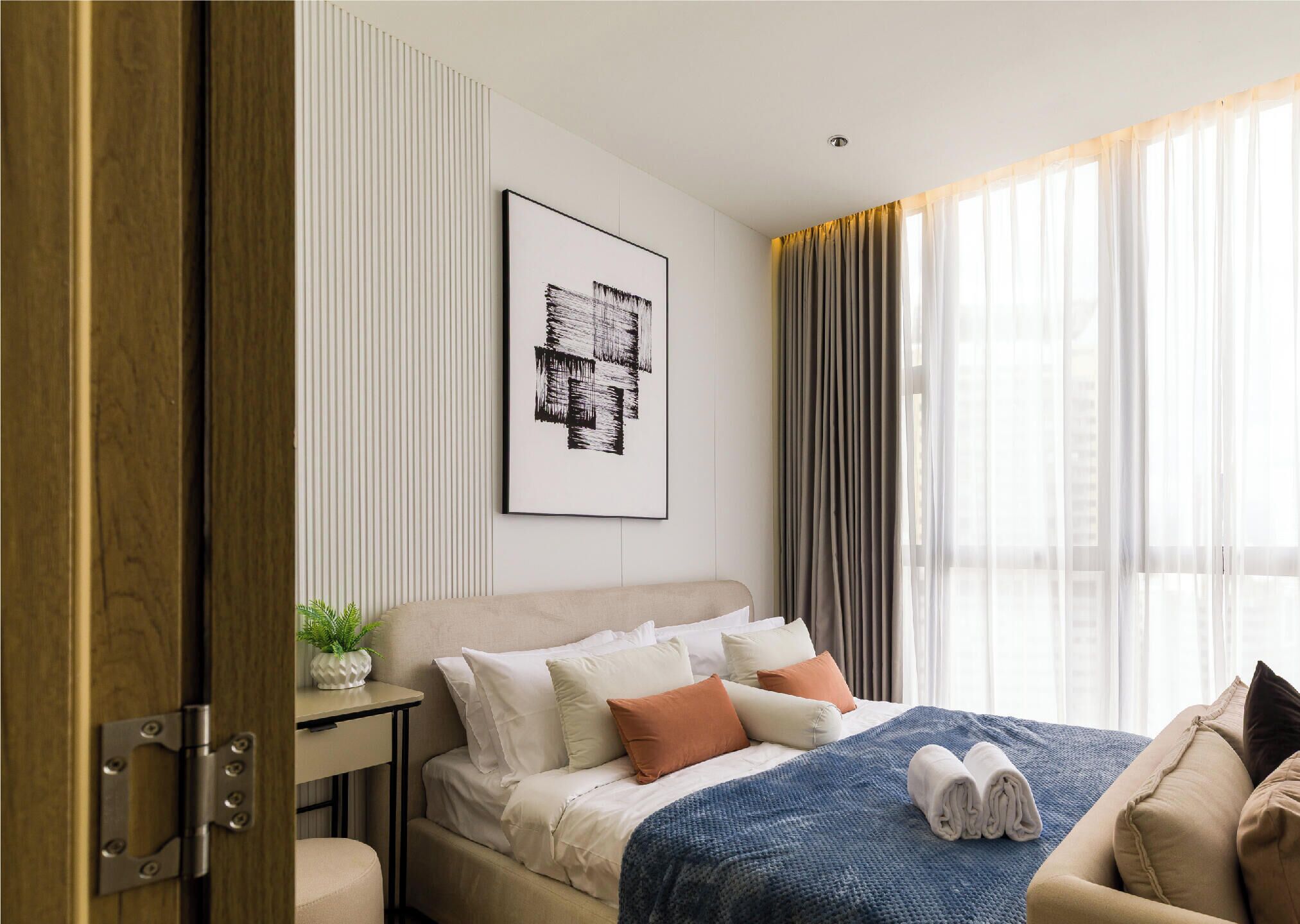 Una habitación de hotel moderna con cama, una mesita de noche con una planta, una obra de arte enmarcada en la pared y una ventana con cortinas.
