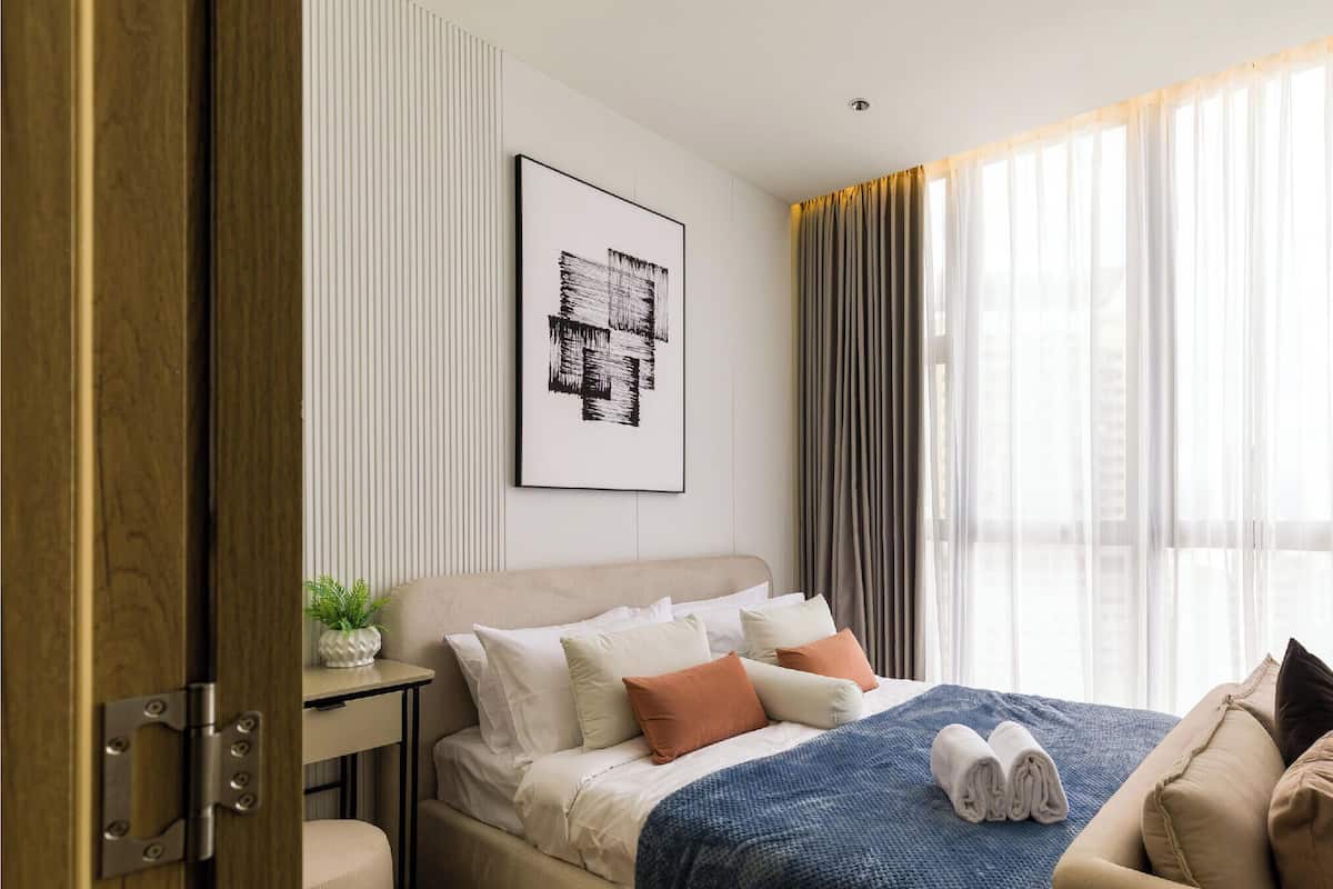 Ein modernes Hotelzimmer mit einem Bett, einem Nachttisch mit Pflanze, einem gerahmten Kunstwerk an der Wand und einem Fenster mit Vorhängen.