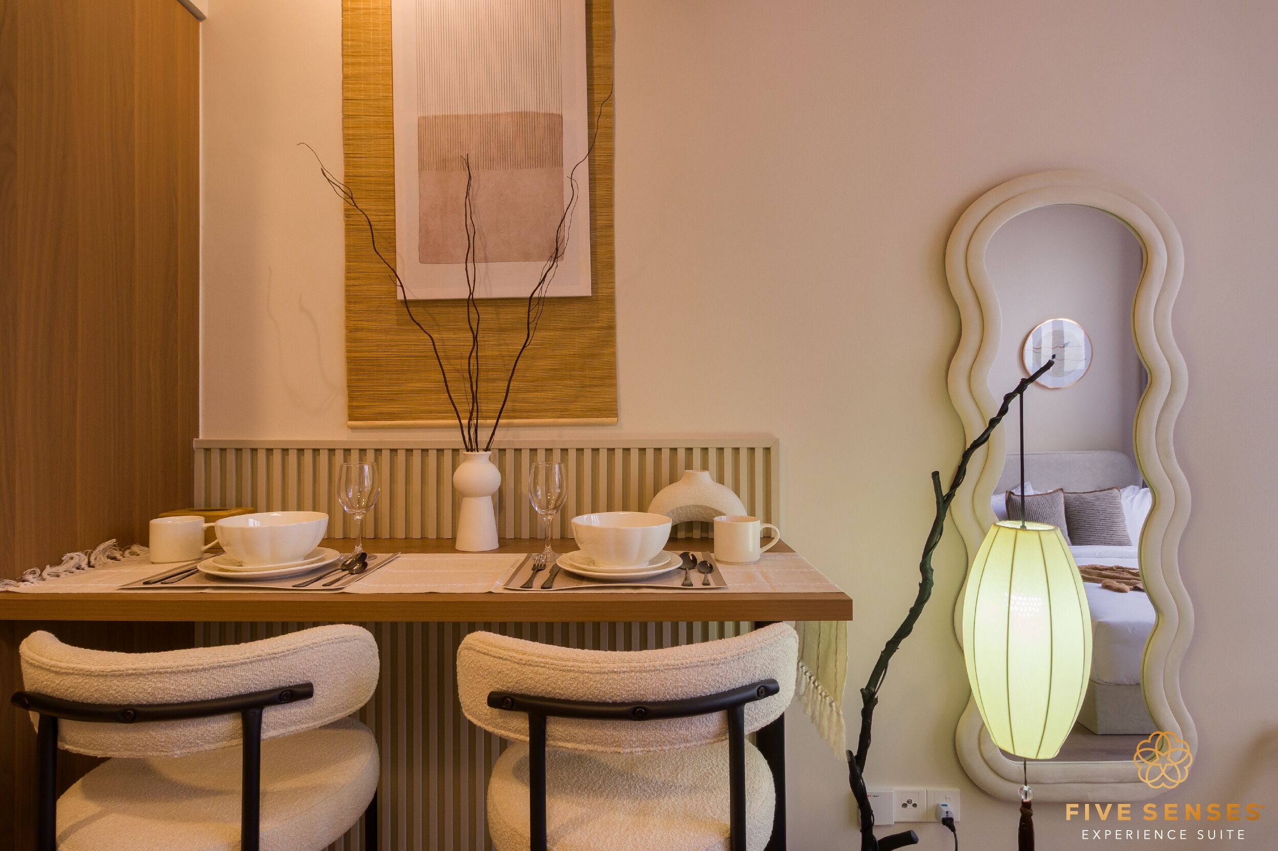 A dining area with a wooden table set for two, a vase with branches, and a wall-mounted mirror with a wavy frame.