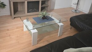 Un salon avec un téléviseur à écran plat posé sur un meuble en bois, une table basse en verre agrémentée d’une plante, un canapé en cuir noir et un tabouret de bar.