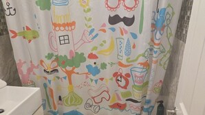 Une salle de bain avec un rideau de douche coloré orné de diverses illustrations ludiques, des toilettes blanches et une baignoire sur le bord de laquelle est accrochée une serviette.