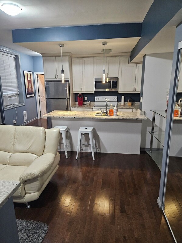 A modern kitchen with white cabinets, a central island, and a built-in oven. There is a beige sofa and two white stools in the living area. - Lovely App Downtown Ottawa 25D (Ottawa)