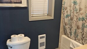 Een badkamer met toilet, een raam met zonwering, een douchegordijn met bloemendesign en een ingelijste foto aan de muur.