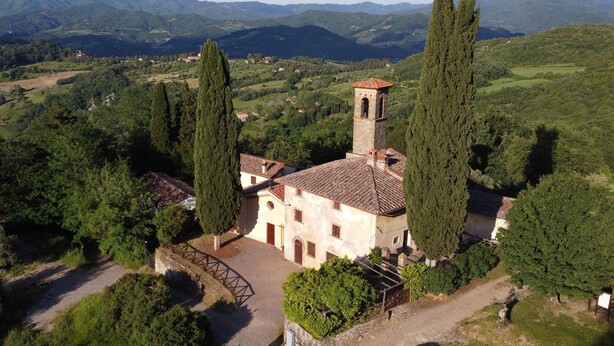 Um edifício histórico com uma torre de sino, rodeado por vegetação exuberante e montanhas ao fundo.