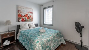 A bedroom with a bed, a nightstand, a lamp, a fan, a window, and a painting on the wall. - Lovely App Downtown Ottawa 211D (Ottawa)