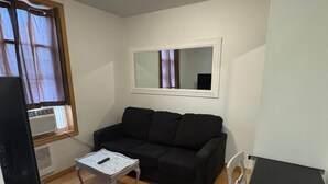 Een kleine, gemeubileerde kamer met een zwarte bank, een klein wit tafeltje, een televisie op een standaard en een raam met gordijnen.