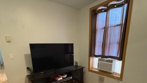 Een woonkamer met een flatscreen televisie op een donker houten kastje, een klein tafeltje met een afstandsbediening, een zwarte bank en een raam met gordijnen.