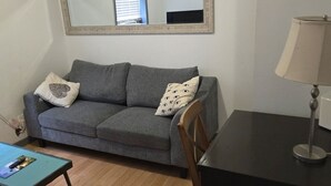Ein kleines, möbliertes Zimmer mit einem grauen Sofa, einem Holzstuhl, einem kleinen Tisch und einer Lampe.